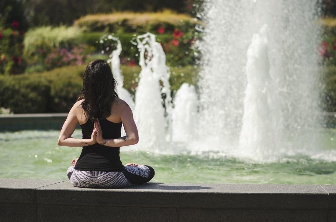 A dark haired woman seated in front of a fountain with her palms pressed together behind her back