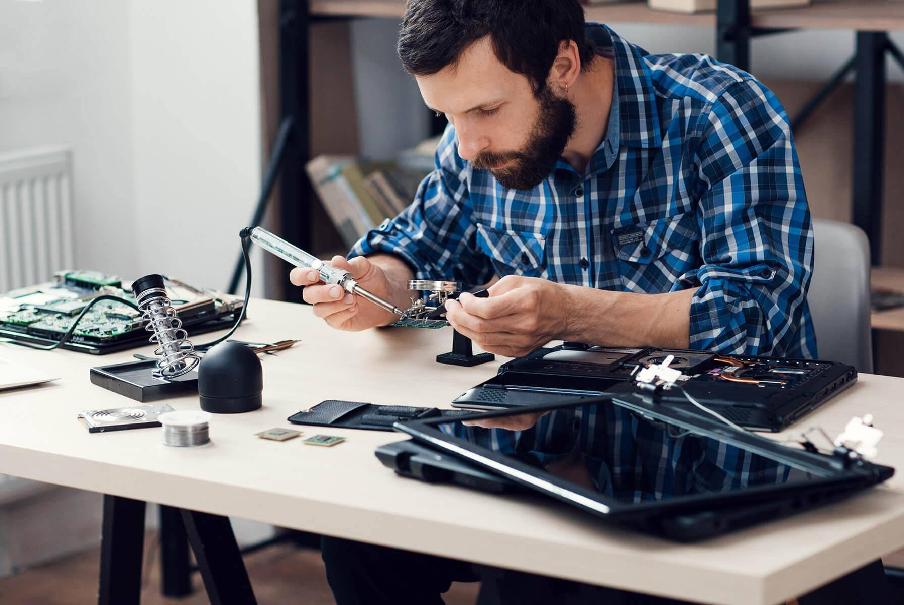 A bearded man using a soldering iron to tinker with electronics piled on a desk