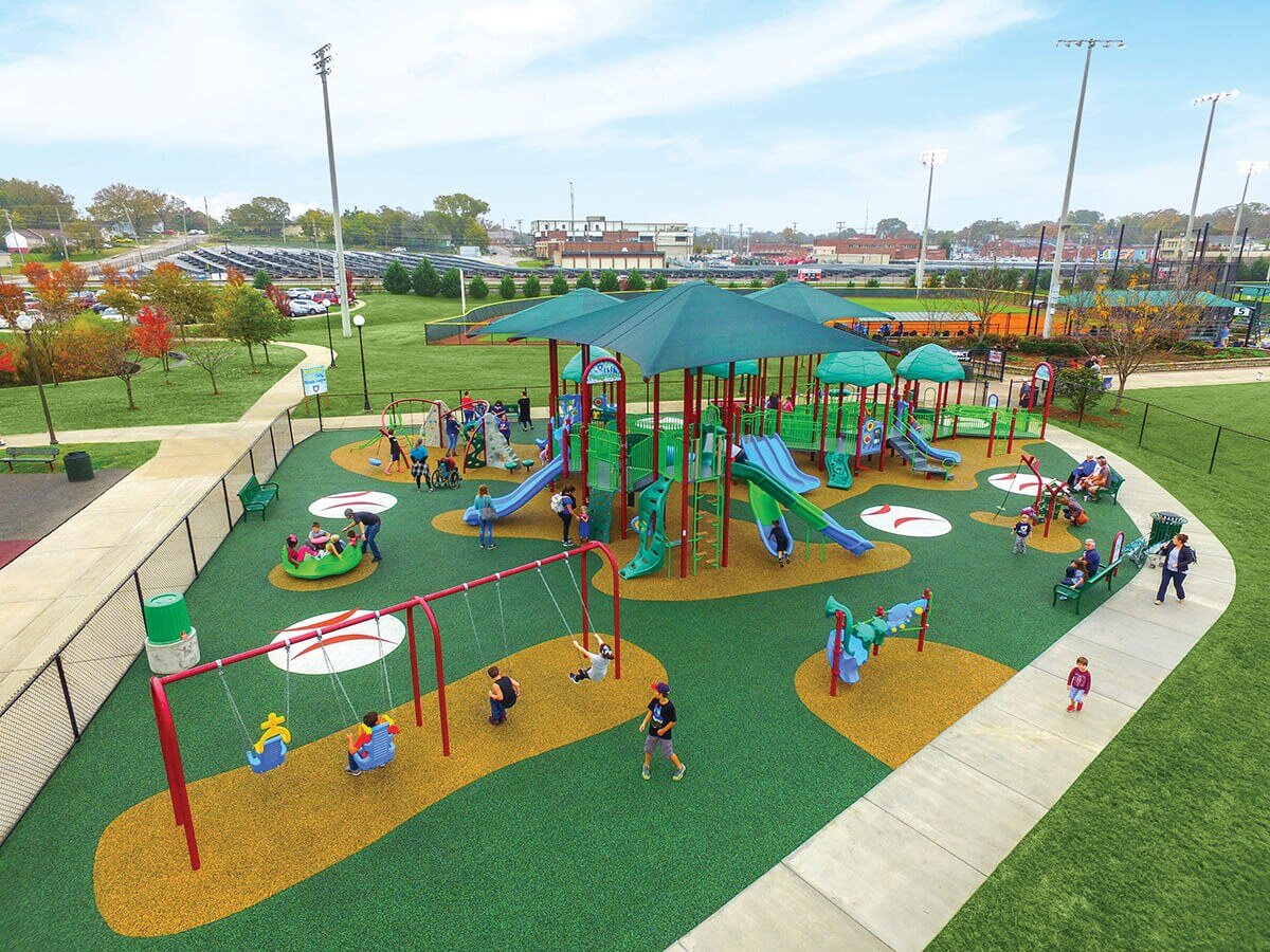 Aerial view of a Miracle League inclusive playground with baseball-themed rubber surfacing, shade-covered slides and swings, and children of all abilities playing.