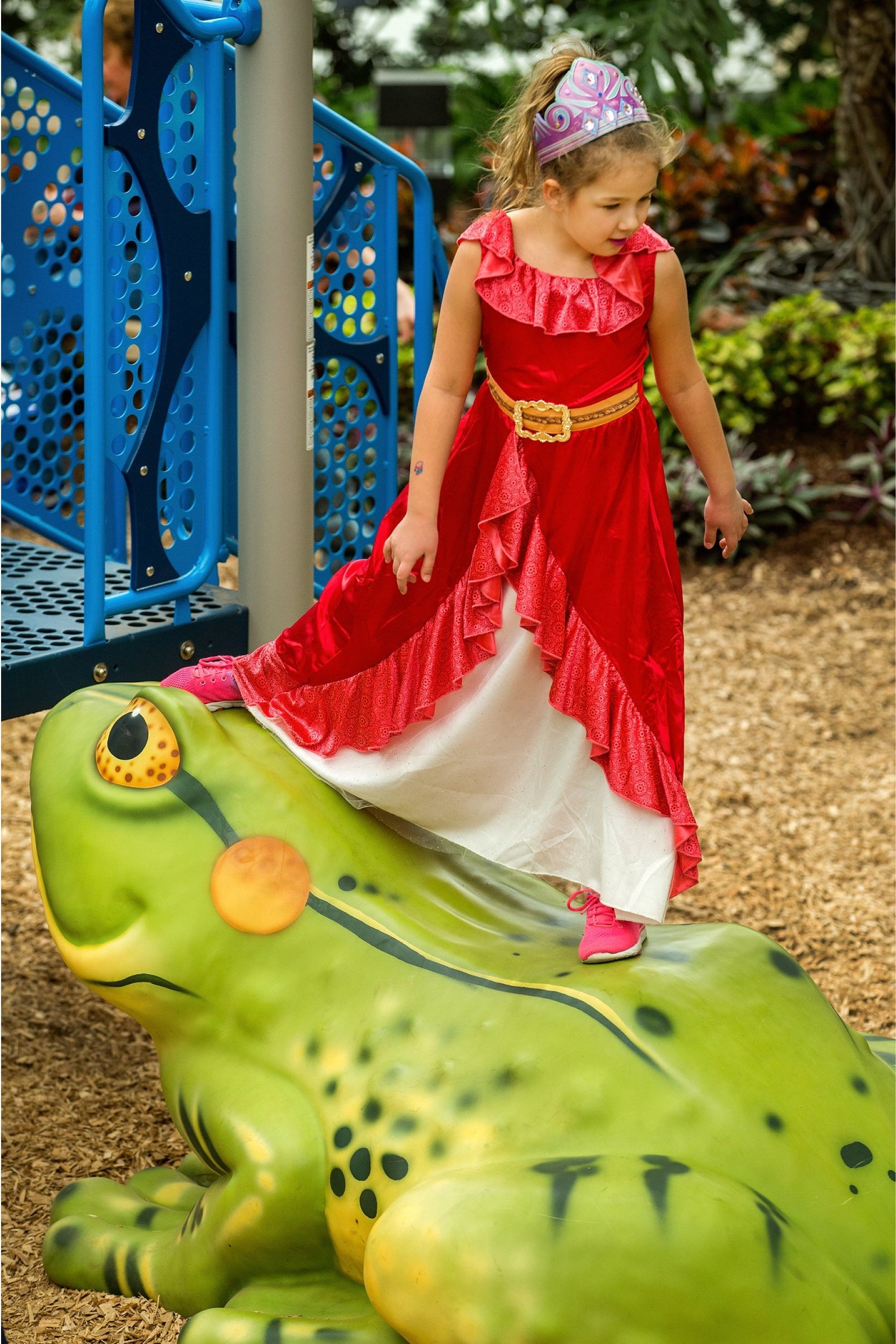 Young girl wearing a tiara and red princess dress balances on a large green frog sculpture at a playground beside blue climbing equipment.