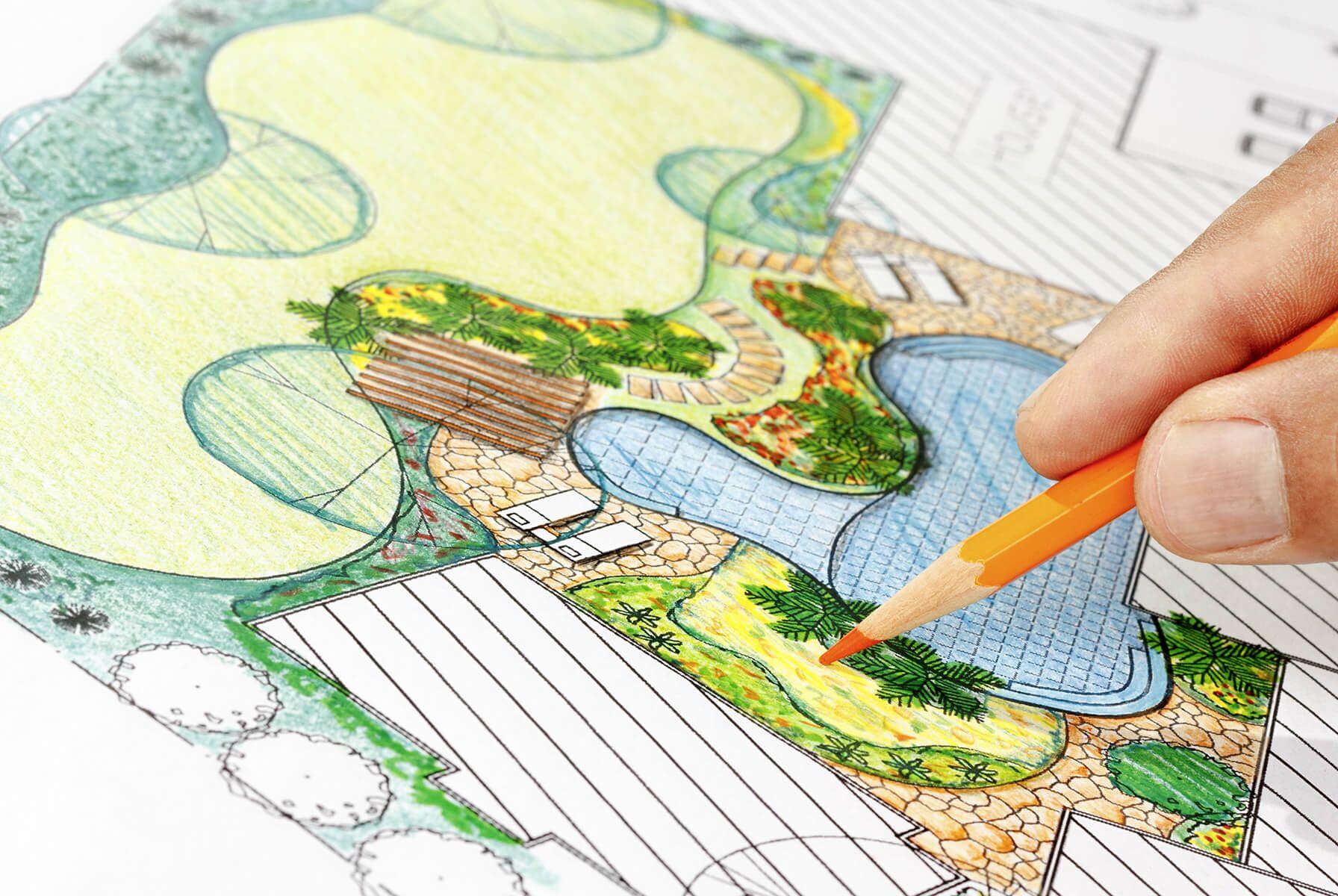 Close-up of a hand using an orange pencil to color a detailed landscape design plan featuring lawns, pathways, plant beds, and a water feature.